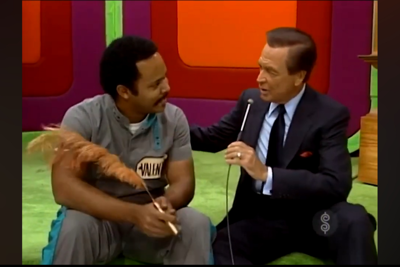 A man holding a feather pen sits on a colorful stage while interacting with a host who is holding a microphone.