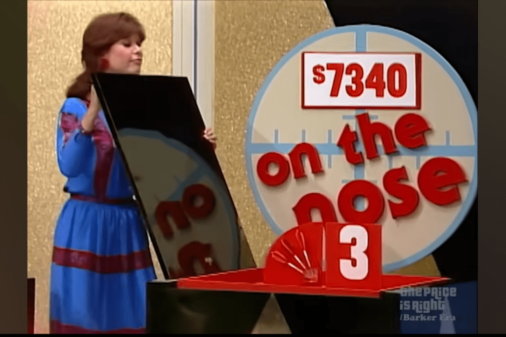 A contestant reveals a black board while standing next to a sign displaying the price 40 with the words 'on the nose' on it.