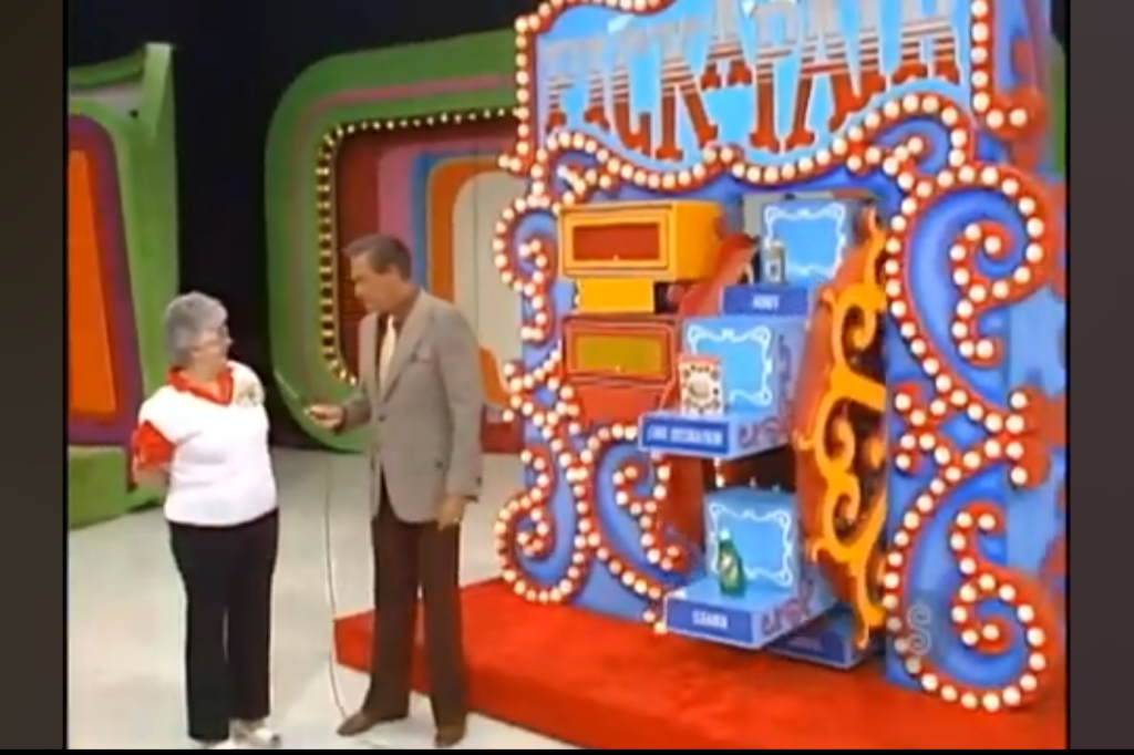 A game show host standing next to a contestant, both smiling in front of a brightly colored game board that features the title 'Pick a Pair' with illuminated lights.