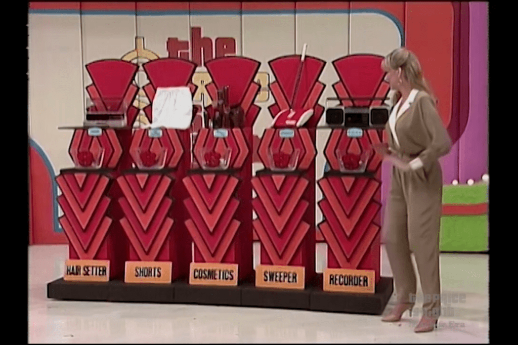 A game show model stands beside a display featuring five small prizes labeled 'Hair Setter,' 'Shorts,' 'Cosmetics,' 'Sweeper,' and 'Recorder.' Each prize is accompanied by a bowl filled with Barker Silver Dollars.