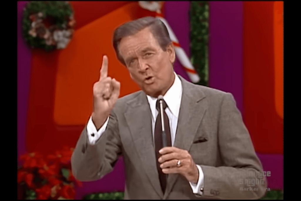 Television host Bob Barker gestures with one finger raised, emphasizing a point while speaking during a game show segment taped around Christmastime.