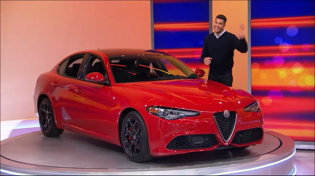 A model presenting a shiny red Alfa Romeo Giulia TI on a game show set, waving to the audience.
