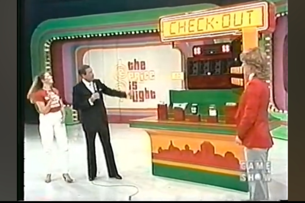 A game show scene from The Price is Right, featuring the 'CHECK-OUT' game setup, with a host gesturing towards the contestant and grocery items displayed on a colorful counter.