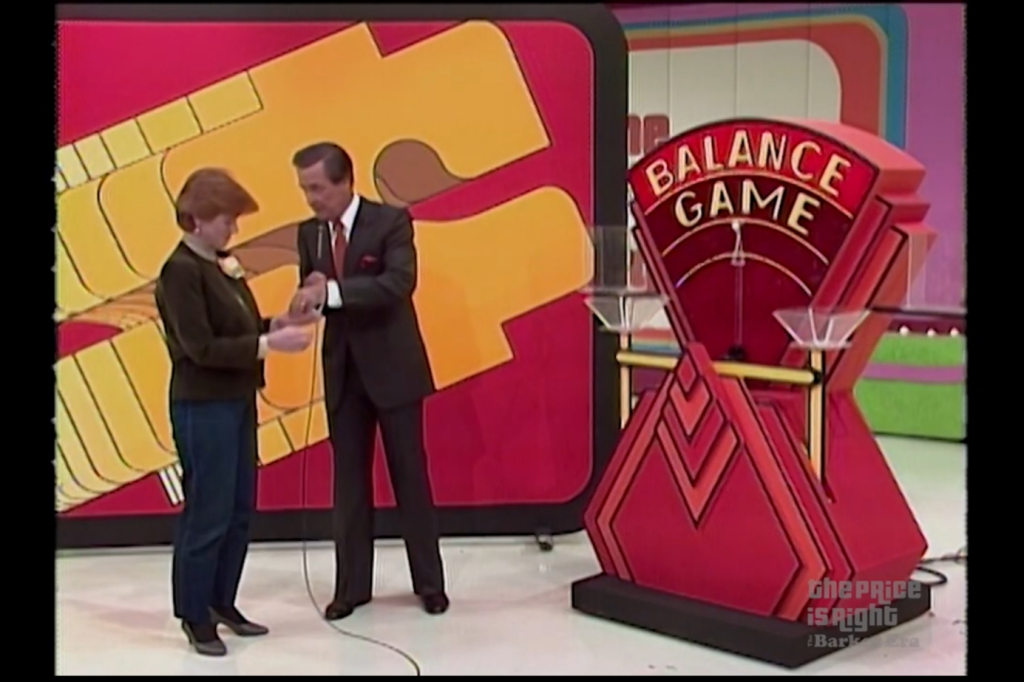 A male host assists a female contestant in the Balance Game, with a large colorful backdrop and a balance scale prominently displayed.