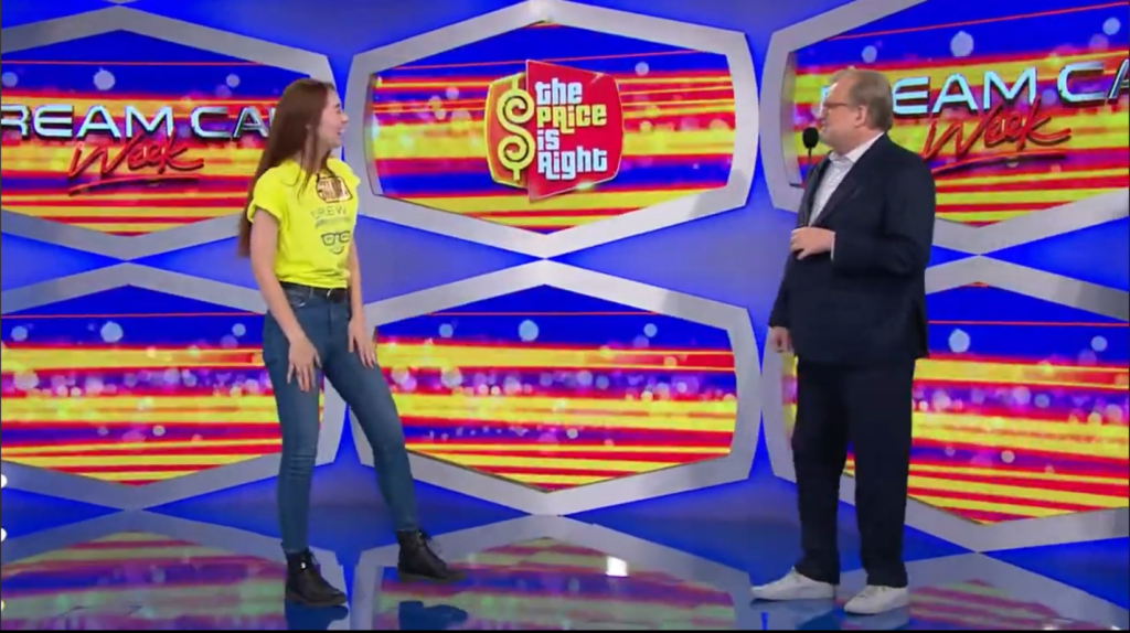 A contestant in a yellow shirt and jeans stands excitedly on stage next to the host during a game show segment of 'The Price is Right'. The vibrant background features colorful graphics promoting 'Dream Car Week'.