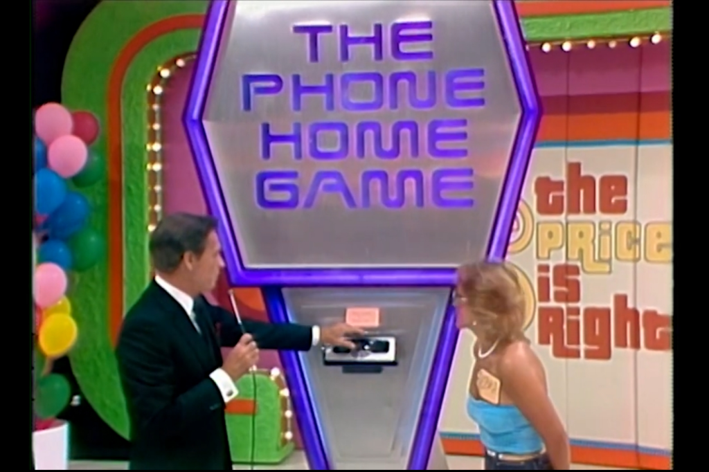Bob Barker explains the rules of 'The Phone Home Game' to a contestant in front of a colorful game board on The Price is Right set.