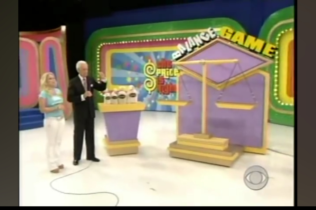 A host stands with a contestant next to the Balance Game setup, featuring a scale and small prizes displayed on a podium.
