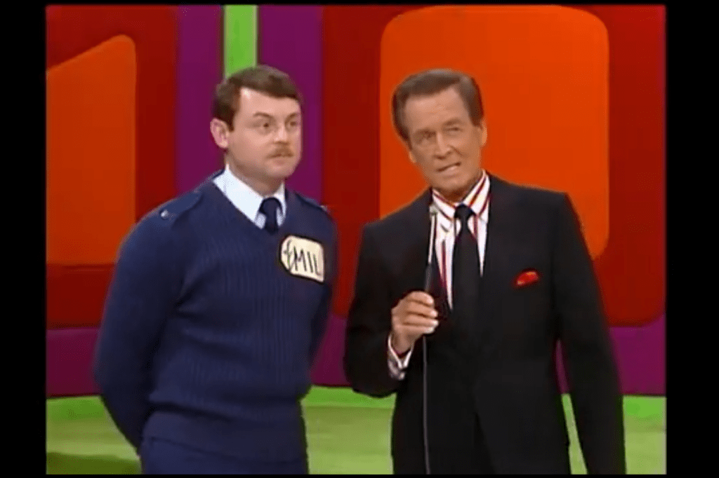 A contestant in a blue uniform stands next to a host in a black suit and striped shirt, both smiling while participating in the game show Balance Game.