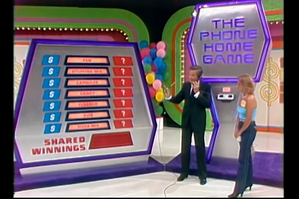 A game show host stands next to a female contestant, explaining the elements of 'The Phone Home Game,' which displays grocery items and potential cash winnings on a game board.