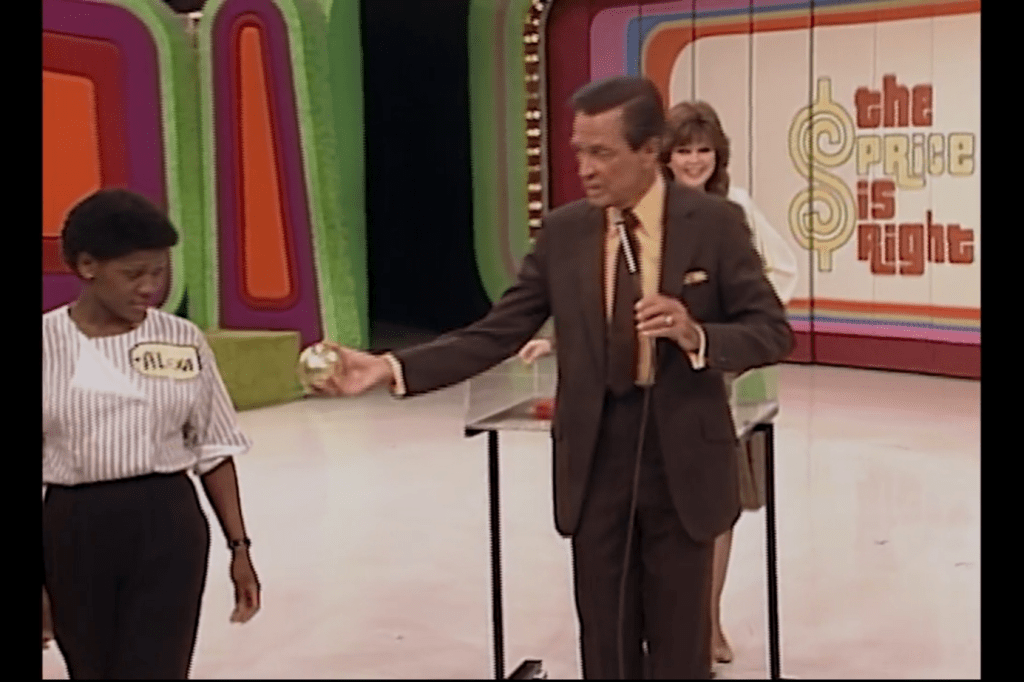 A contestant named Alisa is shown a shiny golden ball by host Bob Barker on the set of 'The Price is Right', while a female model stands in the background.
