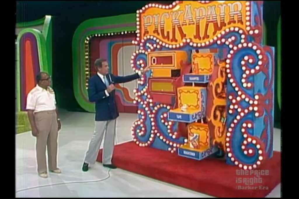 A game show host points to the 'Pick a Pair' game board while a contestant stands nearby, both on a colorful set with various decorative lights and a Ferris Wheel.
