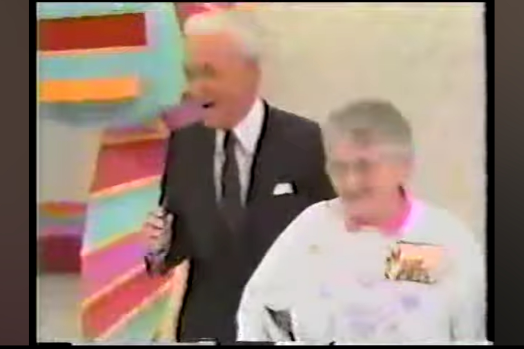 A contestant and host smile and engage during a game segment on The Price is Right, with colorful set pieces in the background.