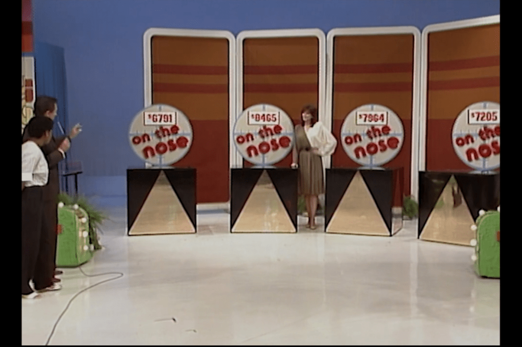 A contestant stands on stage of a game show playing On the Nose, looking at four stands displaying different car prices. The game has a brightly colored backdrop, and the contestant is being guided by a host.