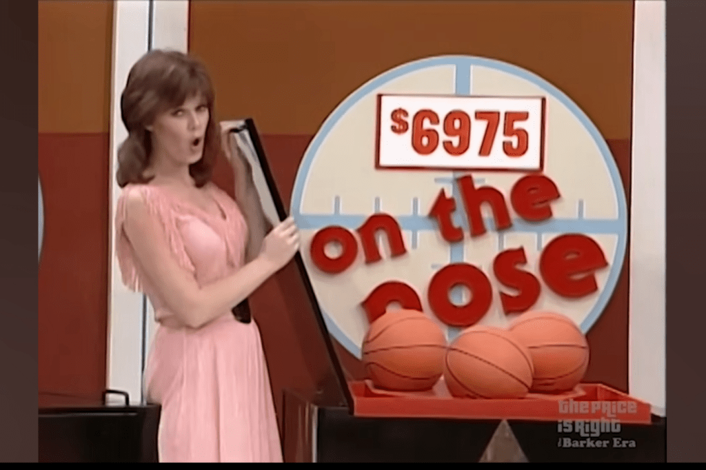A female model in a pink dress stands beside a display showing the game title 'On the Nose' and a price of 75, with two basketballs on the table.