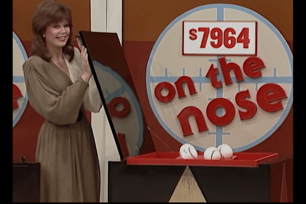 A female contestant smiles while standing next to a display featuring the phrase 'on the nose' and the price 64, along with baseballs in a red container.