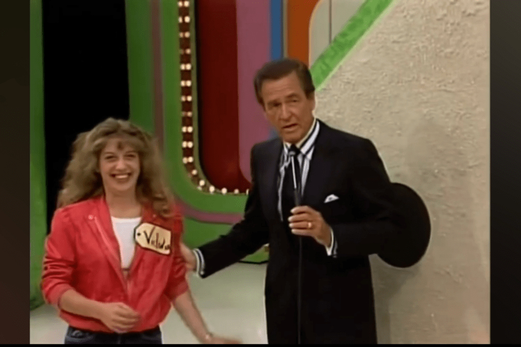 A contestant smiling while standing next to the host of a game show, with colorful set backgrounds.