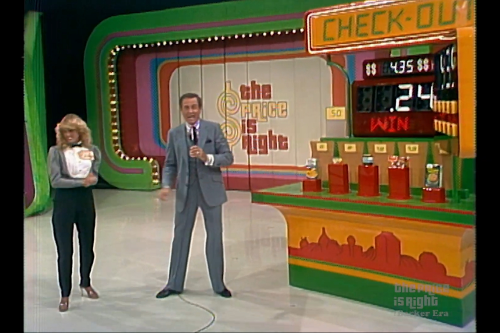 A host stands beside a female contestant on the set of The Price is Right, in front of the Check-Out game display featuring a colorful backdrop and pricing information.