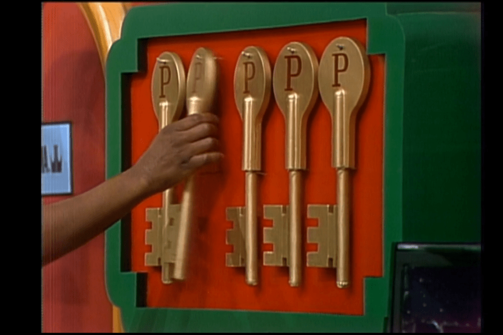 A contestant's hand reaches for one of five golden keys on a colorful display board, featuring three locks below.