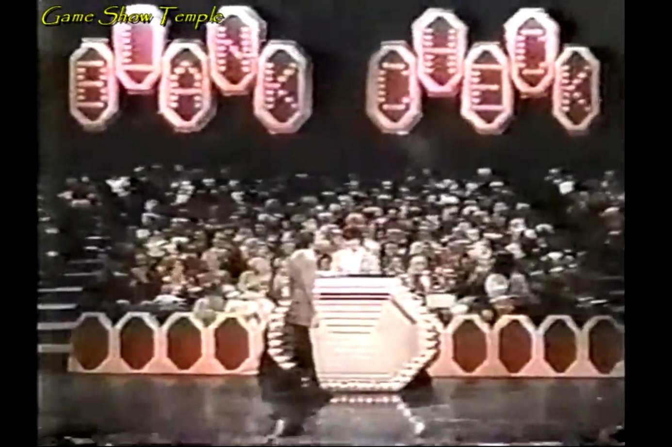 A colorful game show set featuring the title 'BLANK CHECK' illuminated in bright lights, with a host and contestant standing at a central podium in front of a cheering audience.