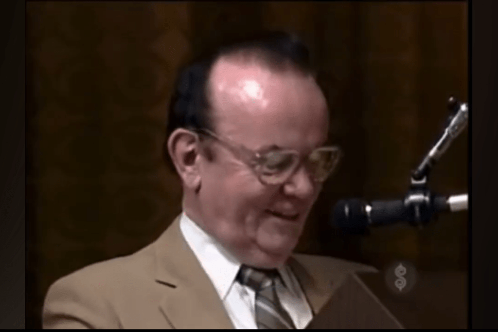 A man wearing glasses and a tan suit smiles while holding a clipboard, standing in front of a microphone.