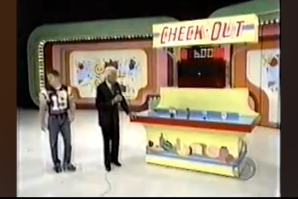Two contestants stand on stage next to a colorful game setup labeled 'CHECK OUT' under bright lights, ready to participate in a pricing game.