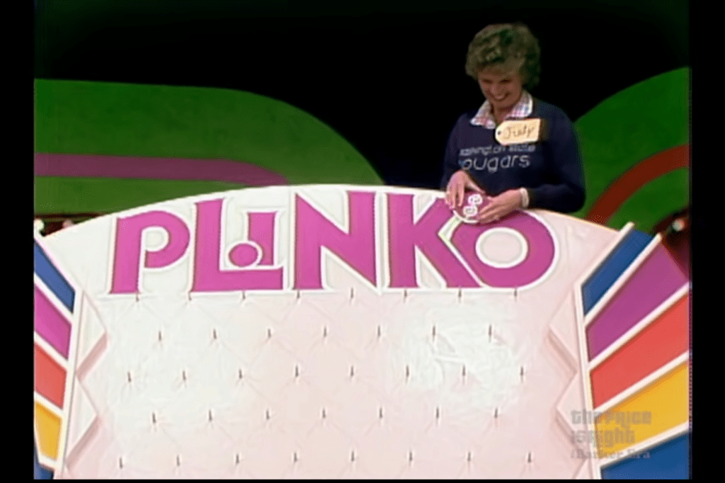 A contestant named Judy smiles while preparing to drop a Plinko chip on a colorful Plinko board, which features the game's name prominently displayed.