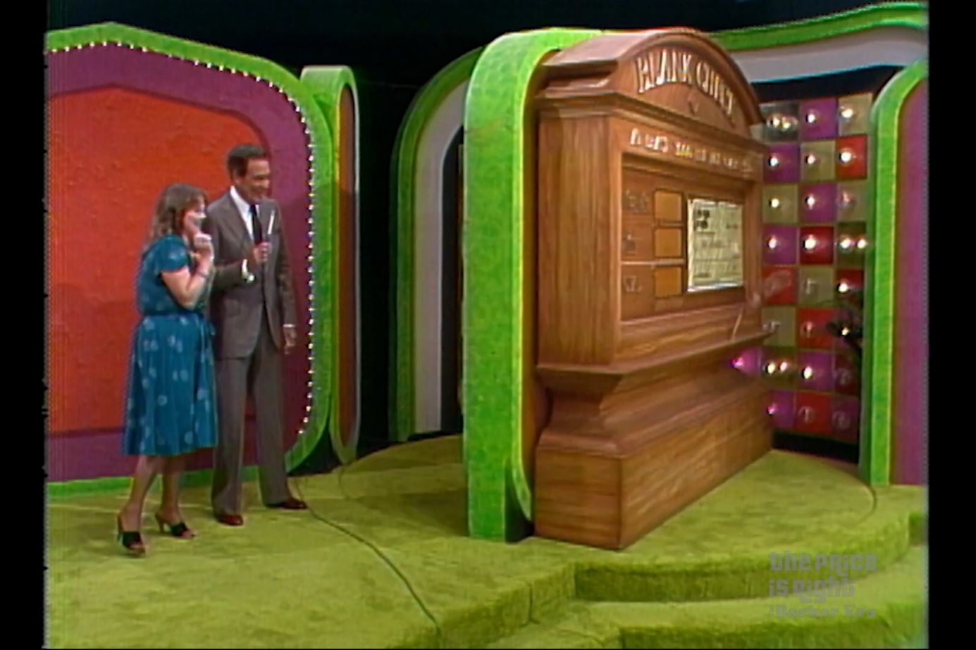 A contestant stands with a host beside a large wooden game display during the 'Check Game' segment on a game show, with a colorful background and lights.