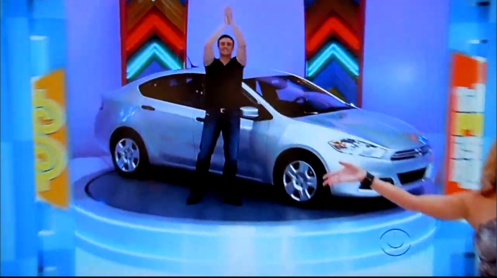 A contestant celebrates with raised hands in front of a silver car displayed on stage during a game show.