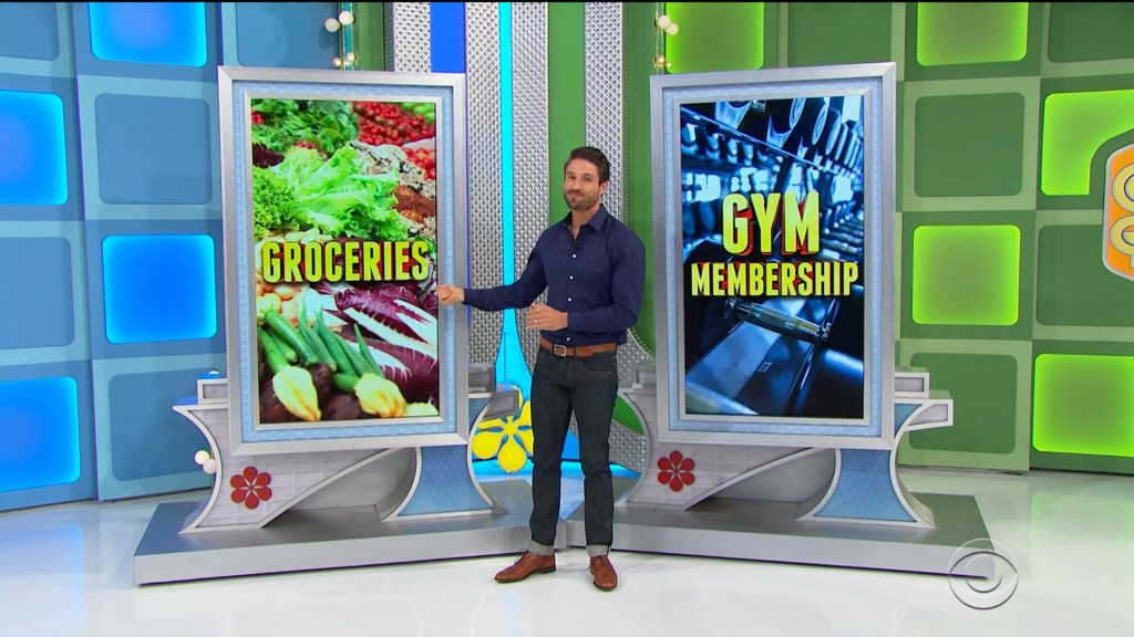 A game show model standing between two large displays featuring the words 'GROCERIES' and 'GYM MEMBERSHIP', with images of fresh produce and gym equipment.