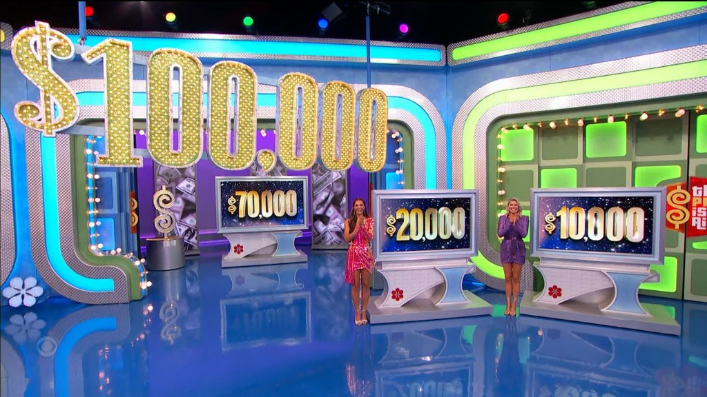 A game show set displaying three prize amounts: ,000, ,000, and ,000, with a large sign above showing 0,000. Two female contestants stand beside the prize displays.