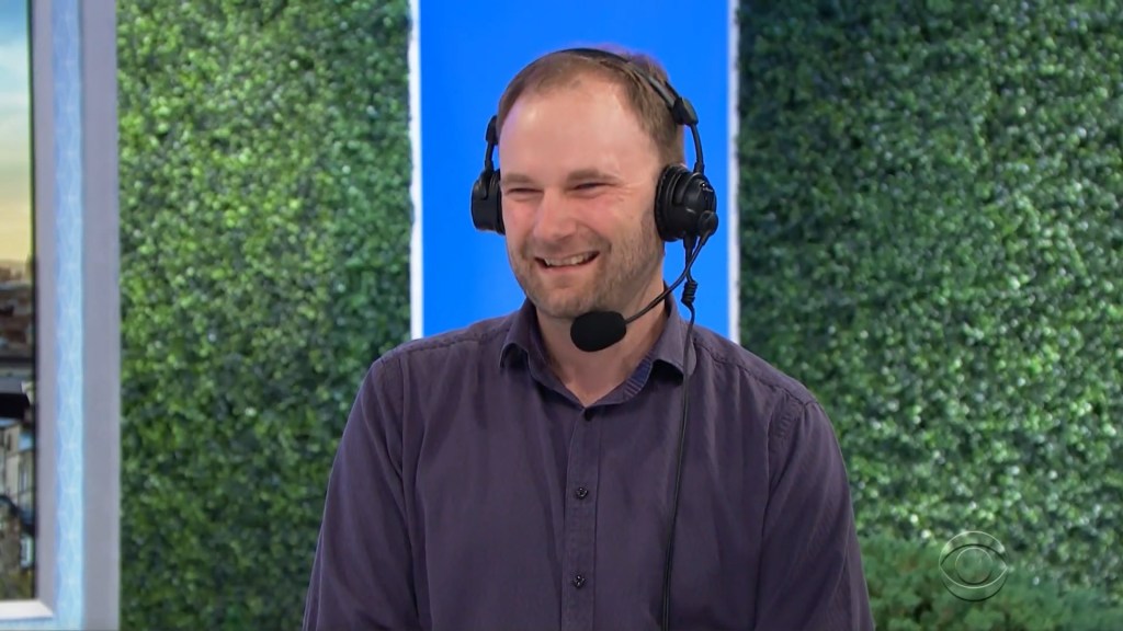 Producer Chris Donnan wearing headphones smiles while playing the Pick a Pair game on a colorful set with a green backdrop.