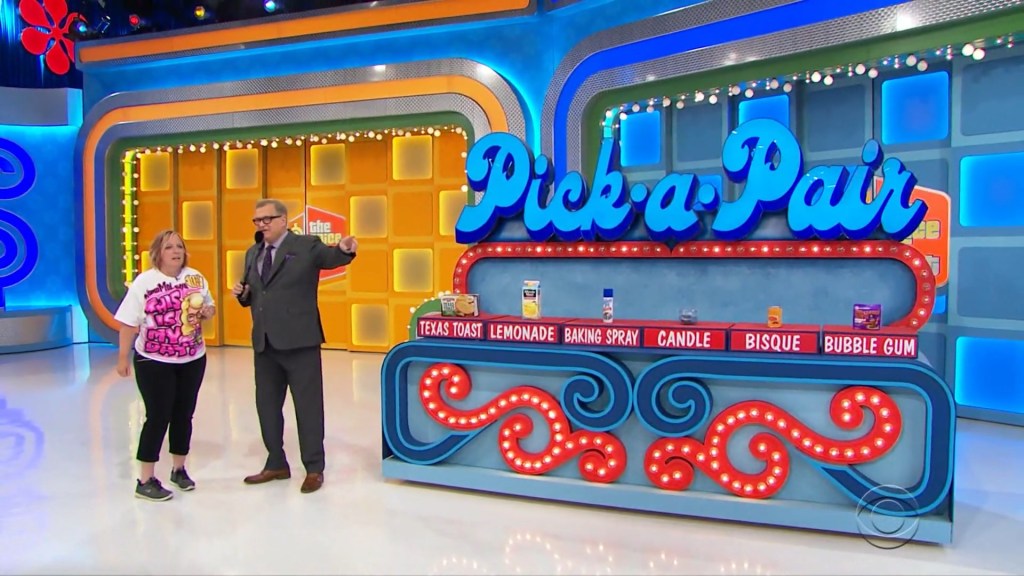 A contestant and host stand in front of the Pick a Pair game set featuring grocery items like Texas Toast, Lemonade, Baking Spray, Candle, Bisque, and Bubble Gum, with a colorful sign above.