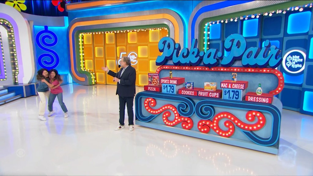 Two contestants celebrate while a host presents the Pick-a-Pair game setup on The Price is Right, featuring grocery items and their prices displayed.