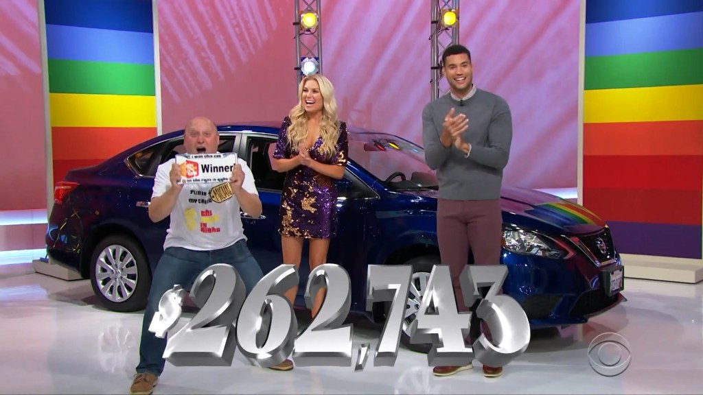 A contestant celebrates holding a 'Winner' sign while showing excitement next to two hosts and a blue car on the set of The Price is Right.