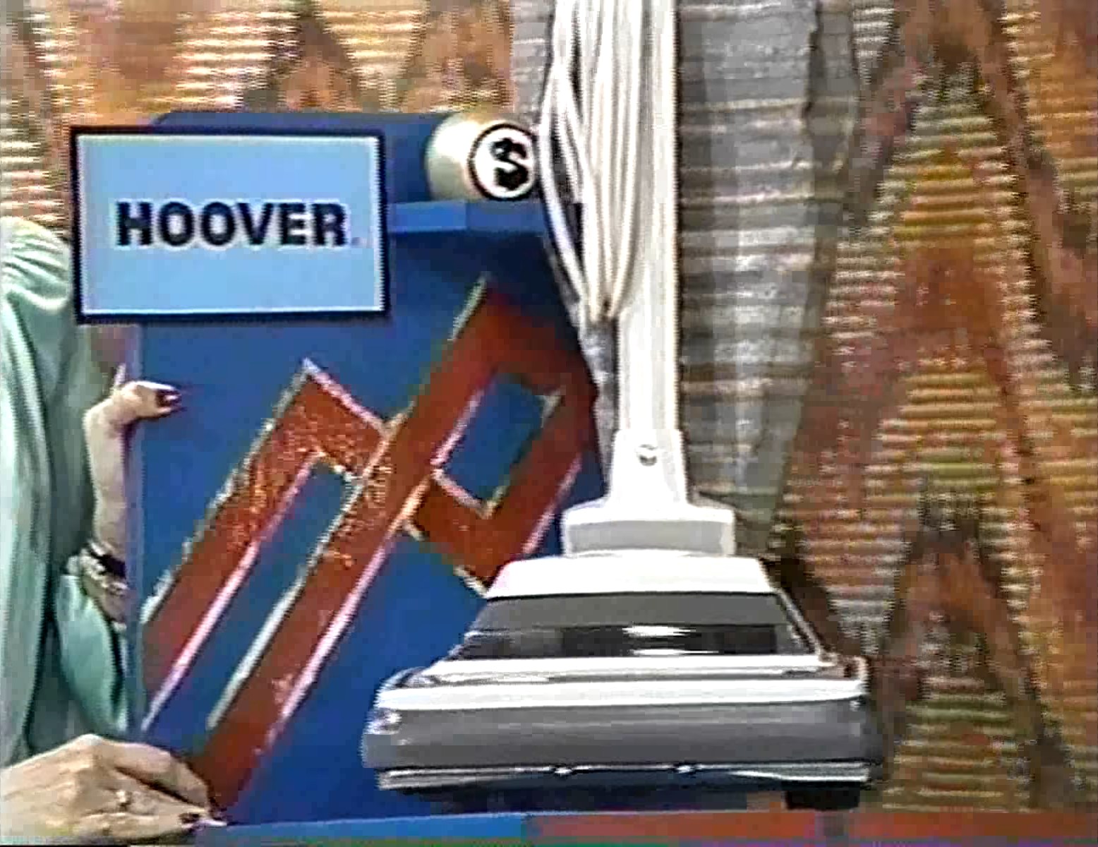 A model holds a sign reading 'HOOVER' next to a vacuum cleaner during a taping of The Price is Right game show, with a patterned backdrop in the Super Ball game.