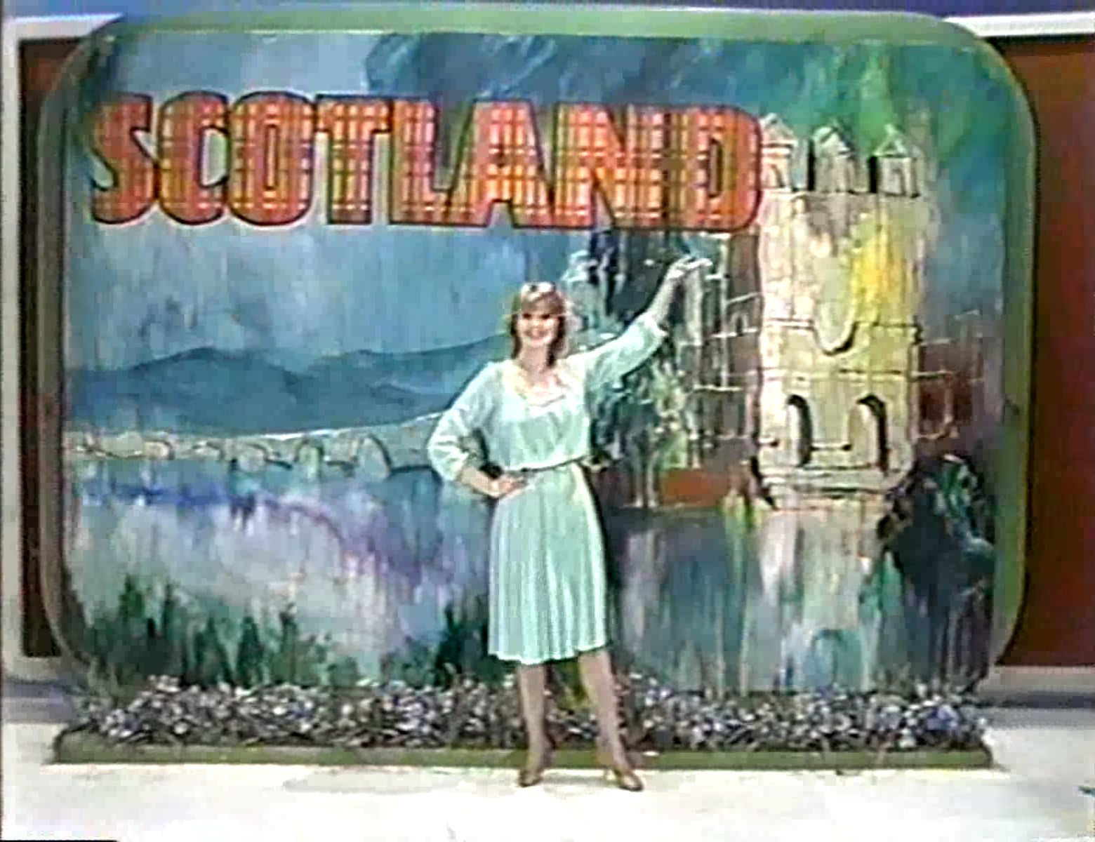 A contestant posing in front of a colorful backdrop depicting a scenic view of Scotland with the word 'SCOTLAND' prominently displayed.