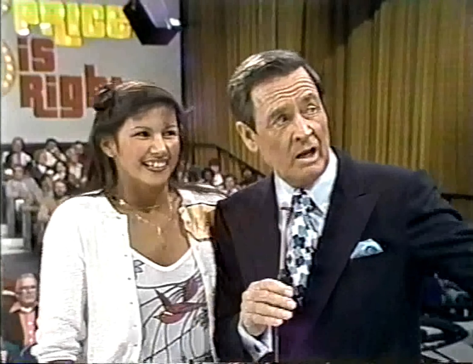 A television host interacting with a contestant on The Price is Right, both smiling, with an audience in the background and a colorful logo of the show visible behind them as they prepare to play Super Ball.