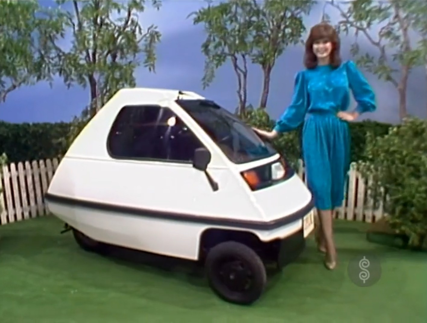 A woman in a blue dress stands next to a small electric car in a green outdoor setting with trees and a white picket fence.