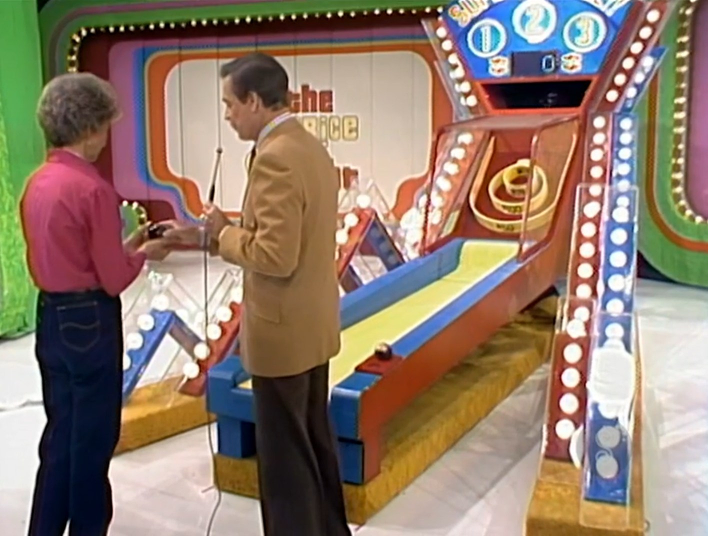 A contestant and host engage at the Super Ball game setup, featuring a colorful ramp and prizes displayed on the background on The Price is Right.