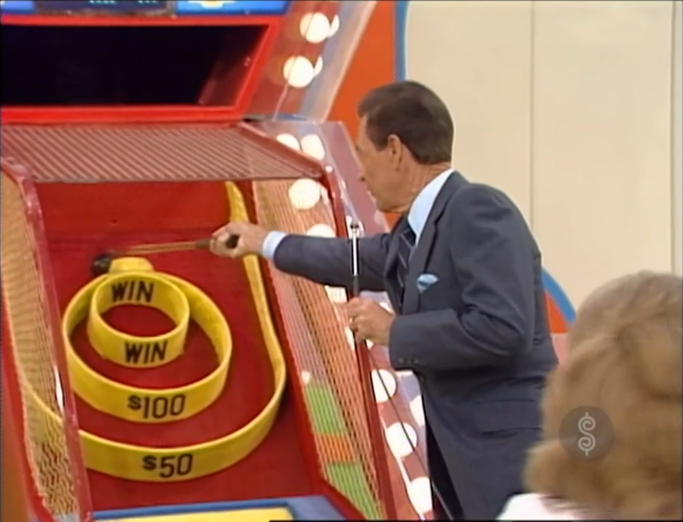 A host demonstrating the Super Ball game on The Price is Right, aiming a ball down a ramp with three circular targets labeled 'WIN', '0', and ''. He is knocking a ball loose with his Plinko Stick.