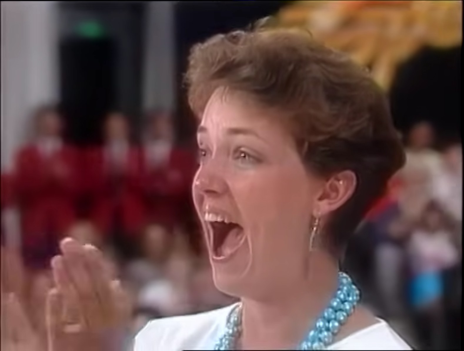 A contestant excitedly reacting with an open mouth and clapping, wearing a pearl necklace, in a game show setting on The Price is Right.