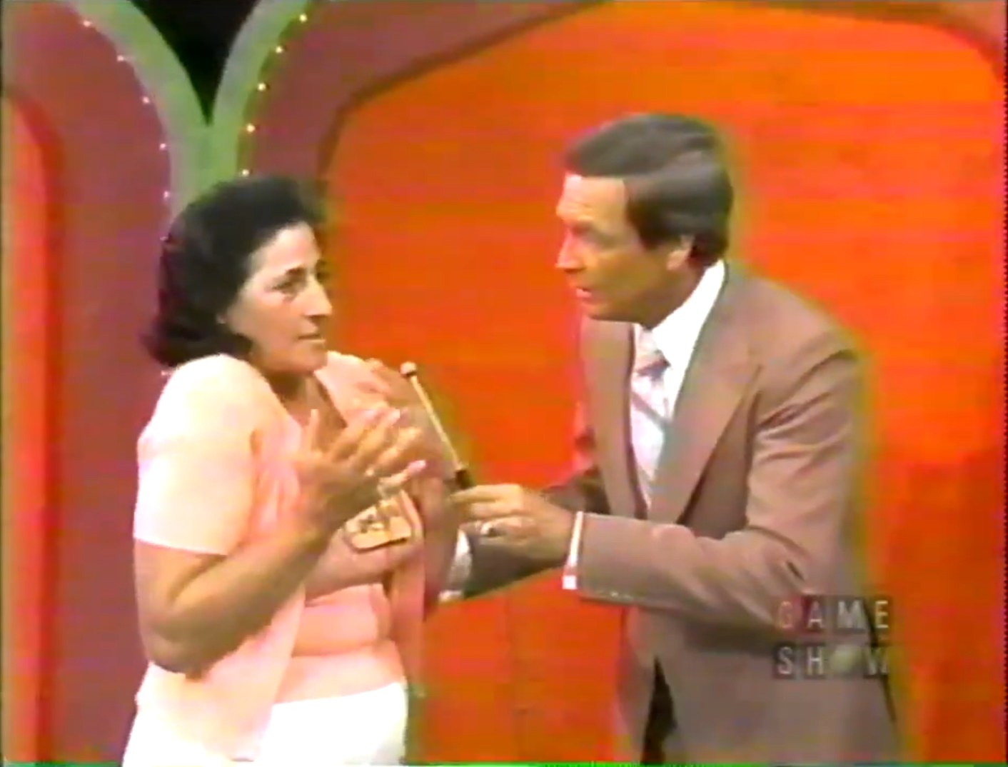 A contestant in a pink outfit expresses surprise or confusion while interacting with the host of a game show, set against a vibrant red background.