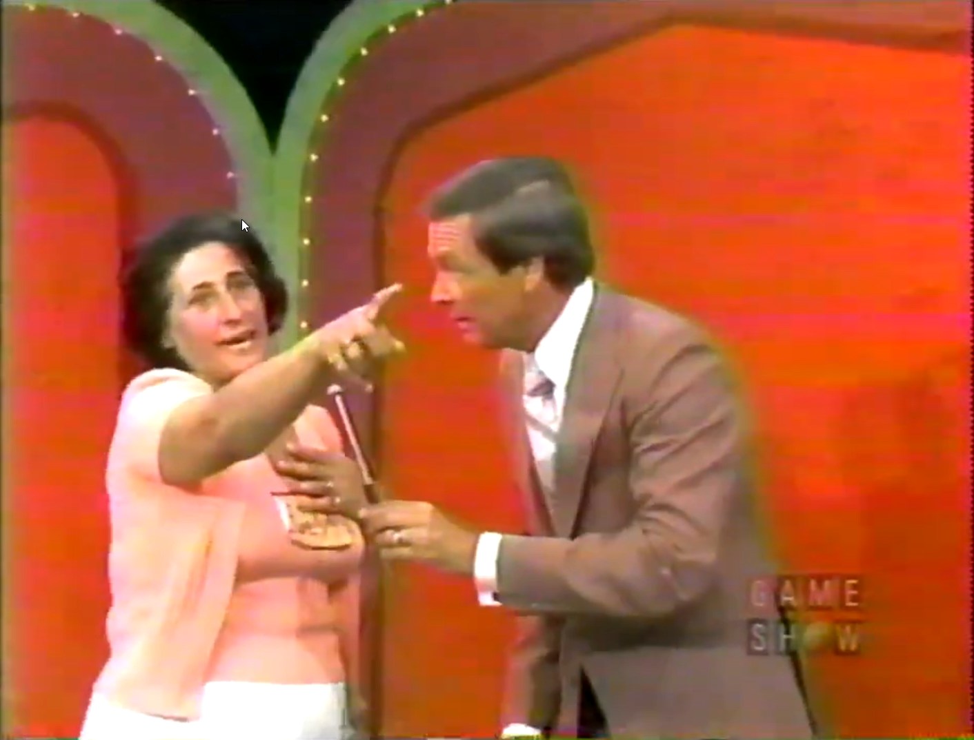 A contestant passionately points while engaging with the host during a game show segment, with bright red curtains in the background.