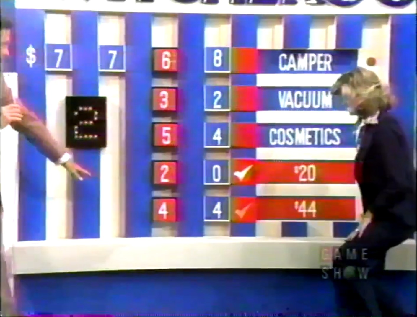 A game show host points at a game board during 'Switcheroo', showing prizes and contestant choices, with numbers displayed and checkmarks indicating correct answers.