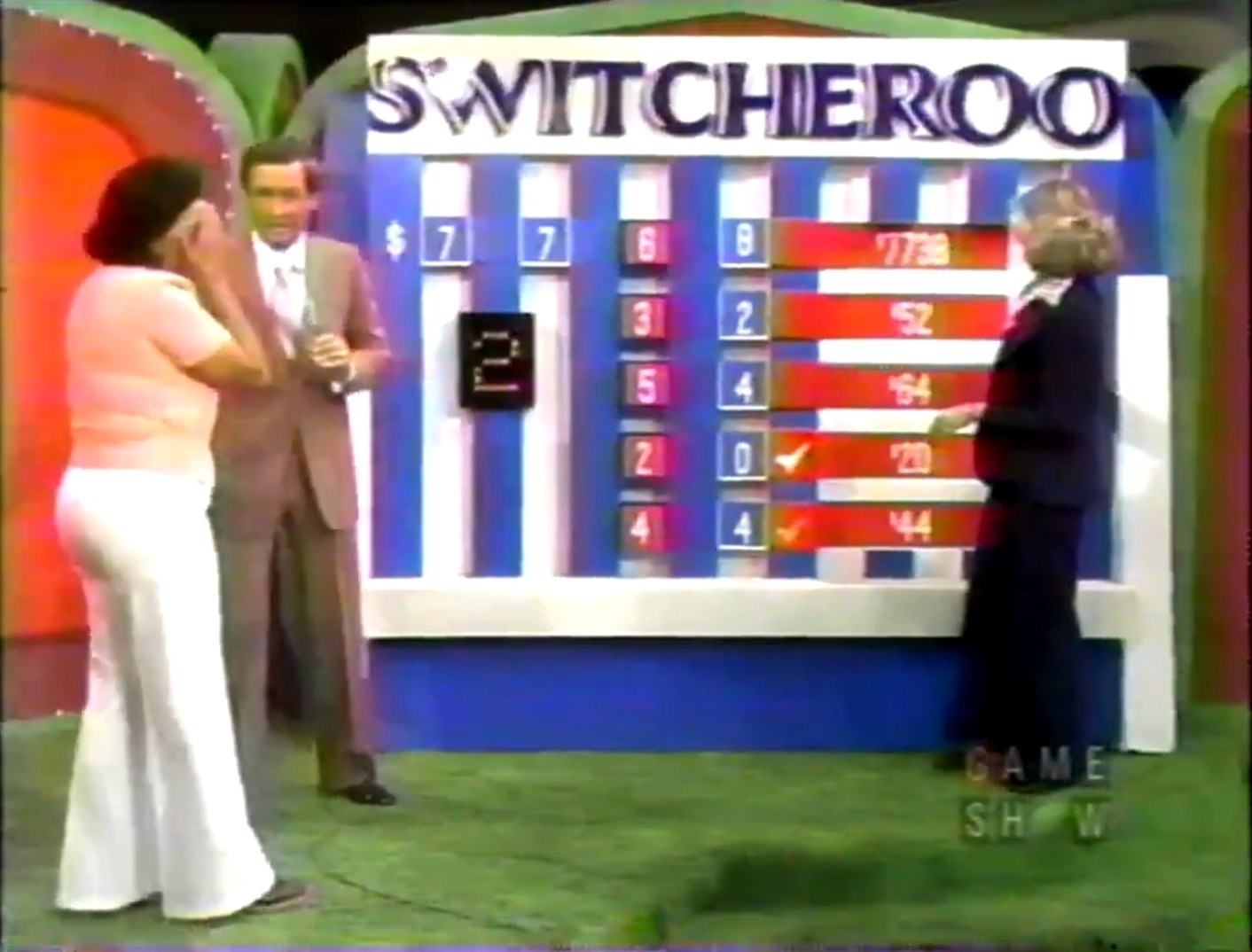 A contestant plays the Switcheroo game on The Price is Right, standing next to the host and a model, with a game board displaying prices and checkmarks.