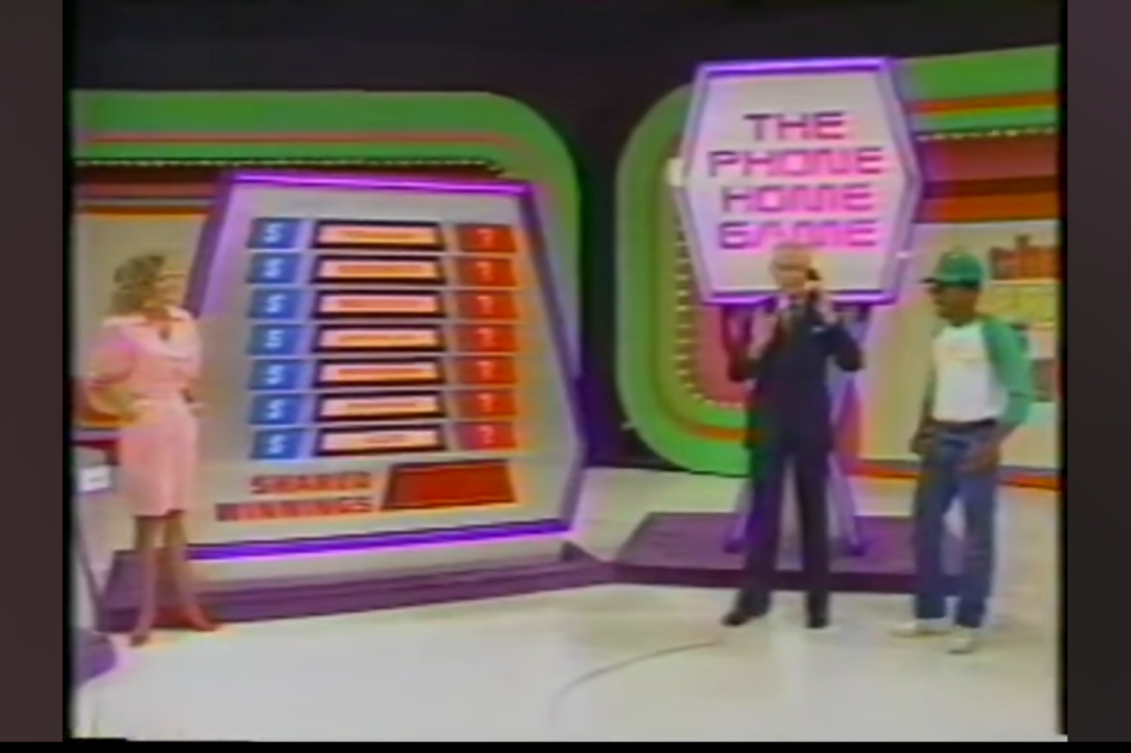 A scene from The Phone Home Game on The Price is Right, featuring a male contestant holding a phone, a host in a suit, and a female co-host in a pink dress next to a colorful game board displaying the game's title and prize structure.