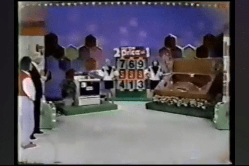 A game show set featuring the '2 for the Price of 1' game, with a contestant and host, alongside a large display showing numbers 769 and 413, and a brown hot tub on stage.