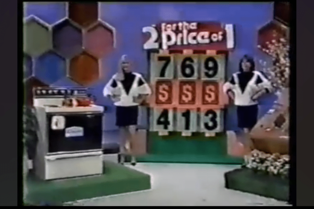 Two female models presenting on the Price is Right with a prize display board reading "2 for the Price of 1" and kitchen range on stage.