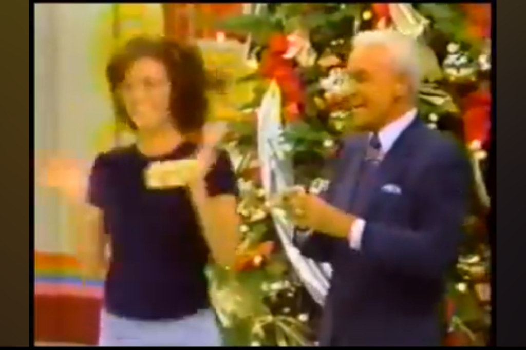 A woman in a black shirt is joyfully clapping her hands, holding a prize, while a smiling Bob Barker in a suit stands beside her against a festive, decorated backdrop with Christmas decorations.