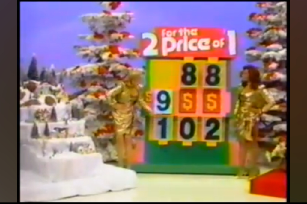 Two models in gold dresses stand beside a colorful game board with the text '2 for the Price of 1' in a festive, winter-themed setting with snow-covered trees and a decorative display.